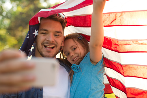 Father and his cute daughter having fun outdoors in a meadow on July 4th. Wearing glasses and American flag. Taking selfie with smart phone. Father holding daughter on his hands. Sun flare, green grass, summer time. Caucasian, blond hair.