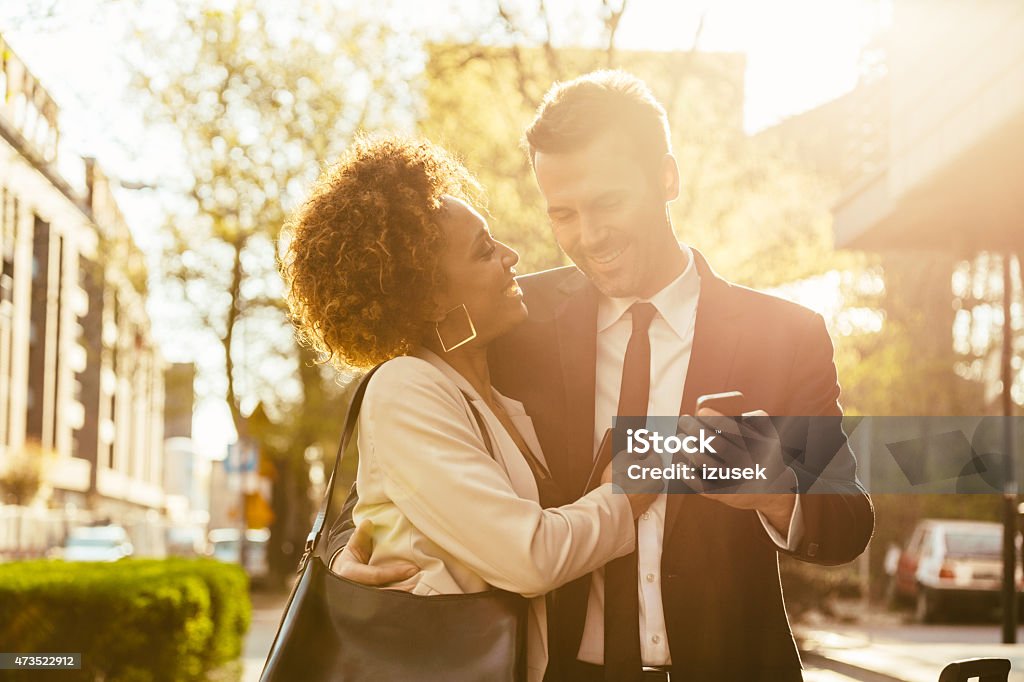 Elegant couple flirting outdoor, man holding smart phone Cheerful caucasian businessman and afro american businesswoman in formal outfits embracing outdoor at sunset. Man holding a smart phone in hand. 2015 Stock Photo