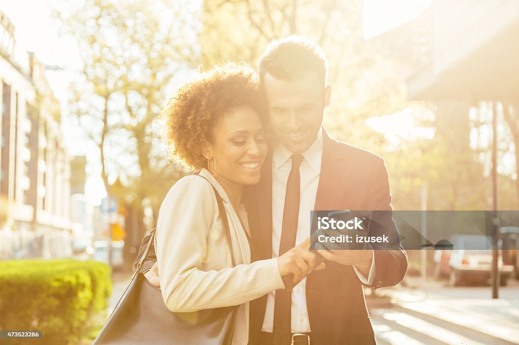 Elegant couple using smart phone outdoor Portrait of cheerful caucasian businessman and afro american businesswoman in formal outfits using a smart phone together outdoor at sunset.  Smart Phone Stock Photo