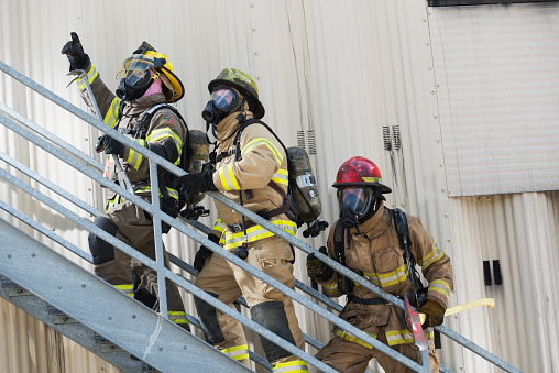 A group of three multiracial firefighters, led by a woman, climbing up a metal staircase on the exterior of an industrial building or warehouse.  She Is holding a crowbar, and has a patch of an American flag on her arm.  They are wearing protective clothing, helmets and oxygen masks.