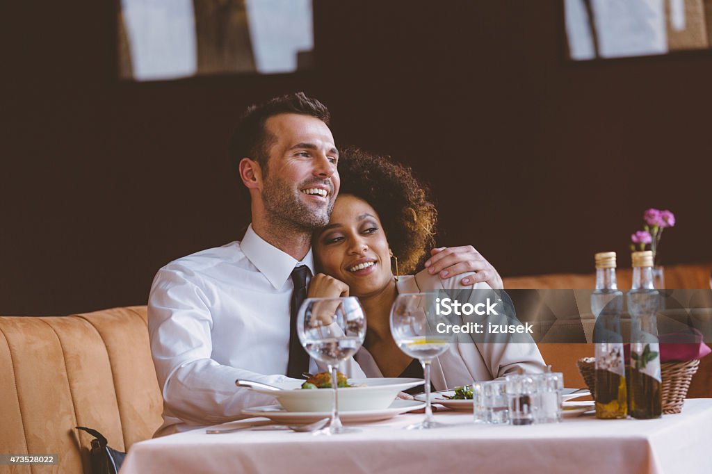 Happy couple at dinner in the restaurant Happy couple - afro american woman and caucasian man in elegant outfits having lunch or dinner in restaurant, sitting at the table and resting after eating. Man embracing her girlfriend. Dinner Stock Photo