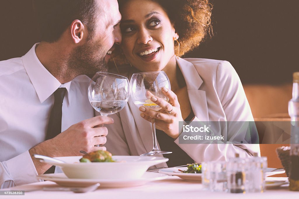 Happy couple flirting at dinner in the restaurant Happy couple - afro american woman and caucasian man in elegant outfits having lunch or dinner in restaurant, sitting at the table and holding wine glasses. Man embracing her girlfriend. 2015 Stock Photo