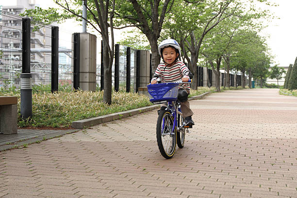 A little boy riding a bicycle in the park with his mother standing behind him