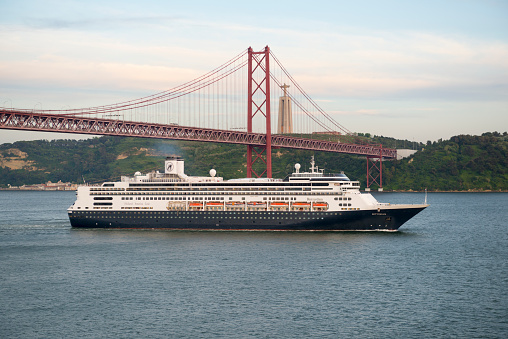 Lisbon, Portugal - May 6, 2014: The MS Rotterdam, a Holland America Line cruise ship, passes under the 25th of April Bridge as it sails out of Lisbon, Portugal. In the distance is the Christ the King statue.