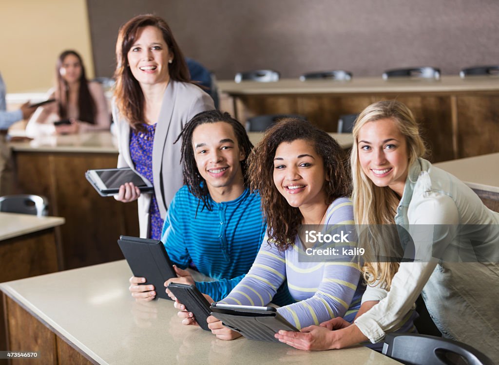 Teacher helping students with digital tablets A teacher with a group of multi-ethnic high school or college students in a classroom.  They are all holding digital tablets, smiling at the camera.  The main focus is on the teenage girl wearing a striped lavender and yellow shirt. High School Student Stock Photo