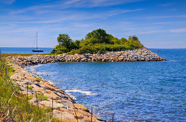 The Knob, rocky shore and waters of the Buzzards Bay, Woods Hole, Falmouth, Cape Cod, USA. The Knob and is a major attraction to the public for its nature walk, birding, marsh, coastal bank and its spectacular views of Quissett Harbor and Buzzards Bay. Green trees, Sailboat and deep blue sky are in background. HDR photorealistic image.