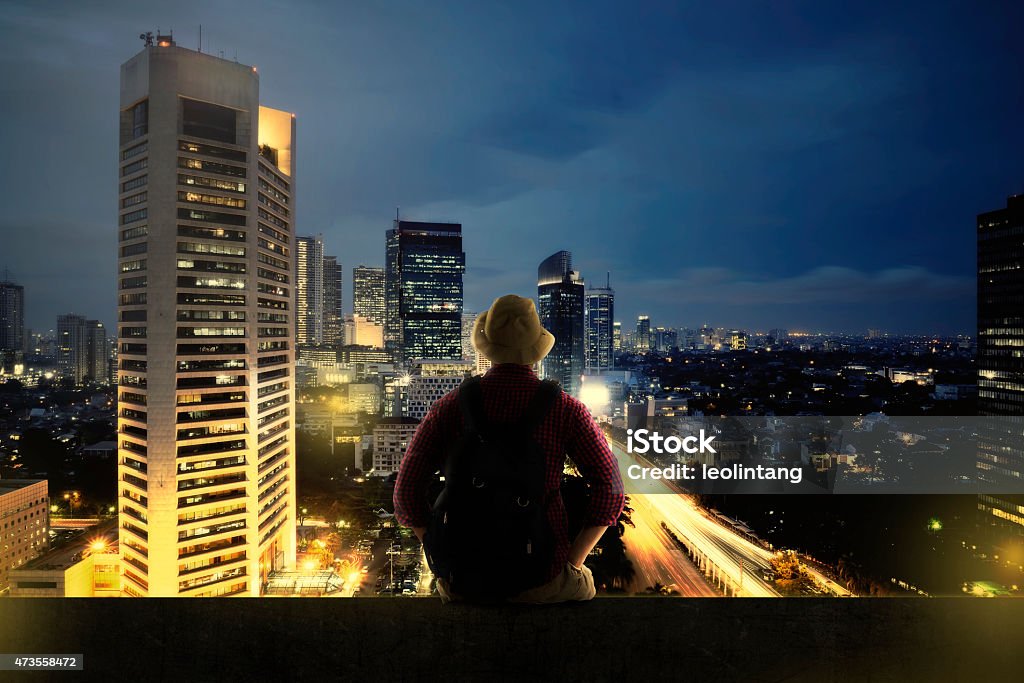 Traveller Sitting On The Top Of Building Traveller man sitting on the top of building looking at the city at night. Business travel concept Jakarta Stock Photo