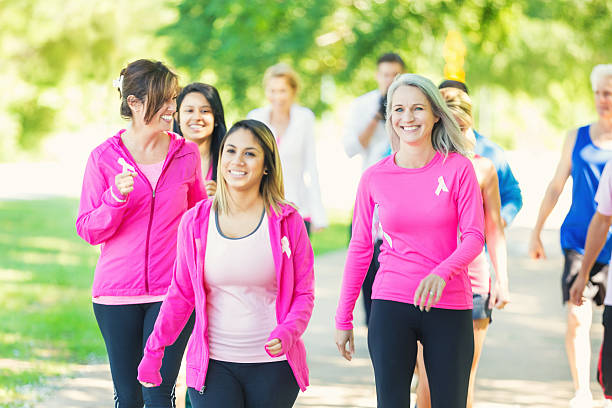 Diverse people are participating in breast cancer awareness race for charity. Young adult Hispanic woman is in the lead, followed by senior and mature Caucasian women and diverse men and women. They are wearing pink athletic clothing and breast cancer awareness ribbons, as they run in marathon or 5k to raise money for breast cancer research charity.