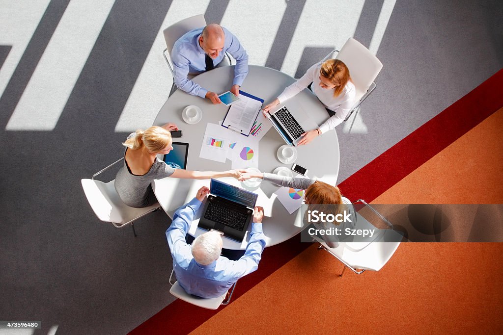 Business meeting Image from above of business people sitting around conference desk and consulting. Businesswomen and businessmen using laptop and digital tablet while working on business plan. Two women shaking hands. High Angle View Stock Photo