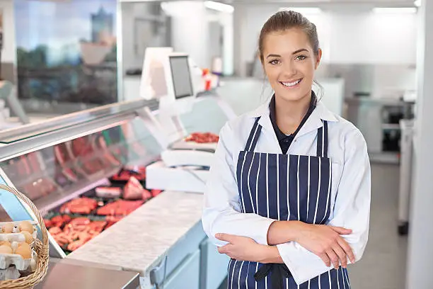 a young female butcher tends the meat counter in a butcher's shop. She is wearing a white coat and striped apron and is smiling to camera. Behind her the meat counter can be seen defocussed .