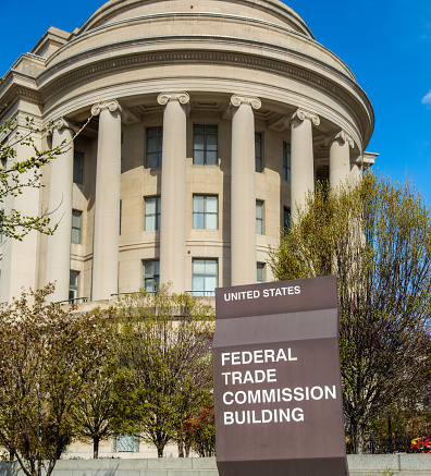 The primary entrance to the Federal Trade Commission Building is at 600 Pennsylvania Avenue NW in Washington DC. Built of Indiana limestone, the round portico is supported by Ionic columns. Leaves on the deciduous trees are just beginning to open under a clear blue sky.