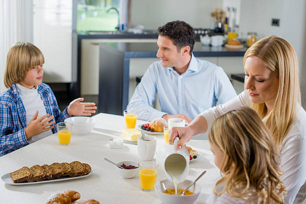 Family having breakfast Father and mother with their children having breakfast at the dining table, mother pouring milk on the cornflakes. Father talking with his son in the background. Glasses of fresh juice and fruits with pancakes on the table. teenager couple child blond hair stock pictures, royalty-free photos & images