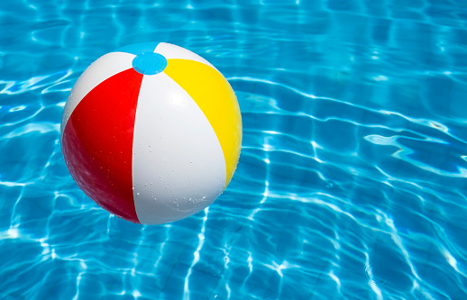 A multi colored beach ball floats on the water surface of a swimming pool. The wave pattern and the bright light blue color of the water gives a feeling of beach party and holiday. With copy space on the right.