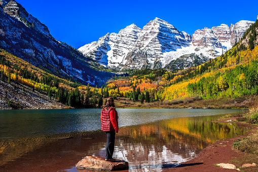 A woman stands on a rock on Maroon Lake admiring the autumnal beauty of the Maroon Bells in a sunny, cloudless day 