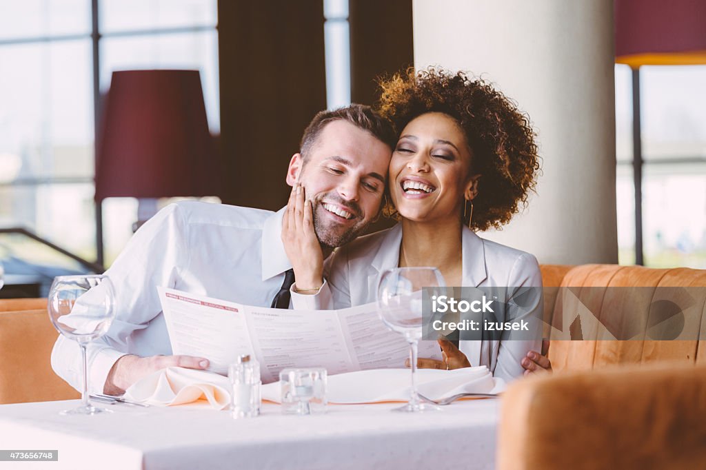 Happy couple at dinner in the restaurant Happy couple - afro american woman and caucasian man in elegant outfits having lunch or dinner in restaurant, flirting at the table. Woman holding menu. Couple - Relationship Stock Photo