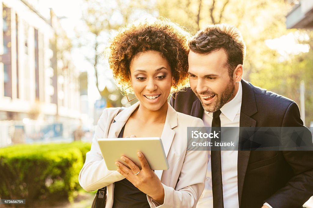 Two business people using digital tablet outdoor at sunset Portrait of cheerful caucasian businessman and afro american businesswoman in formal outfits using a digital tablet together outdoor at sunset.  Businessman Stock Photo