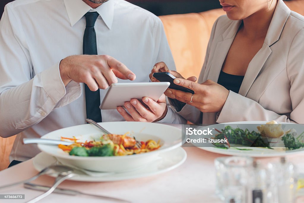 Business people using smart phone and digital tablet at lunch Businesswoman and businessman having lunch or dinner in the restaurant, man using digital tablet and woman using smart phone. Close up of hands. Plates on the table. Unrecognizable person. 2015 Stock Photo