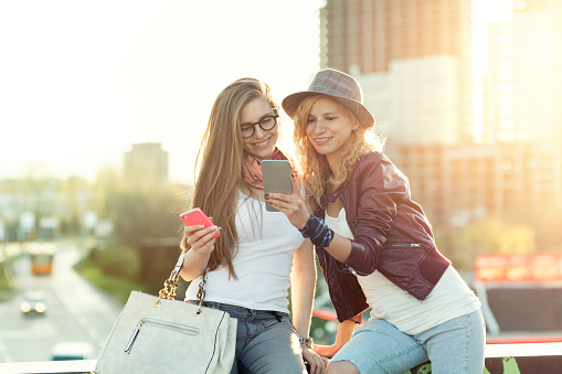 Two young blond girls sitting on a bridge and looking pictures on a smart phone. Casual clothing with jeans and jacket.
