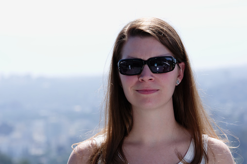 Quarter length portrait of young woman wearing sunglasses and smirking.  Woman is outdoors with city skyline in background.