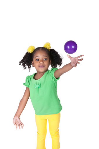 Four year old african american girl is throwing purple plastic ball. She has pigtails with bright yellow hair bows in them. She is wearing a green shirt and yellow pants. She is standing up with arm outstretched. Her mouth is closed.  Shot taken with a Canon 5D Mark 3. rm