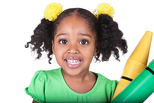 An adorable four year old african american girl is holding two big colored toy crayons. She has pigtails with bright yellow flower hair accessories around them. She is looking at the camera with a toothy smile. Studio shot, isolated on white, taken with a Canon 5D Mark 3 camera. rm