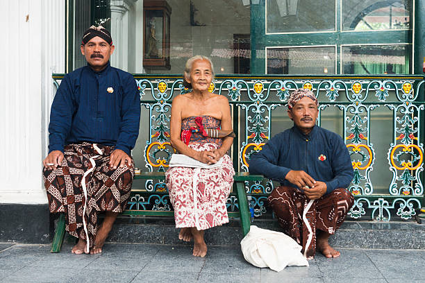 Javanese People Wearing Traditional Clothing at Yogyakarta Palace Indonesia Yogyakarta, Indonesia - April 6, 2015: Three Javanese people wearing traditional clothing sit outside during a break while working at the Sultan's Palace in Java, which is also called the Kraton. The two men wear head cloths and sarongs. The senior woman who sits in the middle wears a light colored sarong. yogyakarta stock pictures, royalty-free photos & images