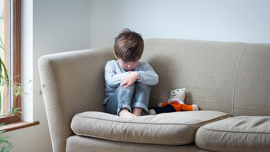 Little boy suffering from child abuse curled up on the sofa with his teddy.