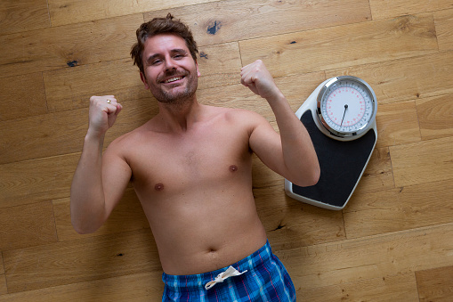 Man lying down next to bathroom scales looking up to camera and shaking his fists in triumph.