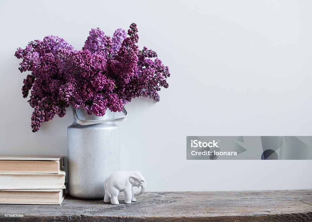 Purple flowers in a vintage milk tin with porcelain elephant Home interior decor, bouquet of lilacs in a vase and books on rustic wooden table, on a white wall background Purple Stock Photo