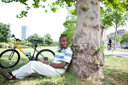 Young man sitting in the park leaning against a tree. His bicycle is next to him and ho holding a mobil phone