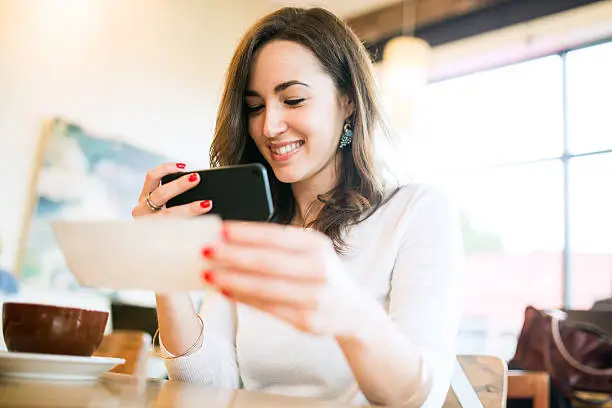 A smiling young woman takes a picture with her smart phone of a check or paycheck for digital electronic depositing, also known as "Remote Deposit Capture".  She sits in a coffee shop, enjoying an espresso latte.  Bright sunlight shines in the windows behind them.  Horizontal image.