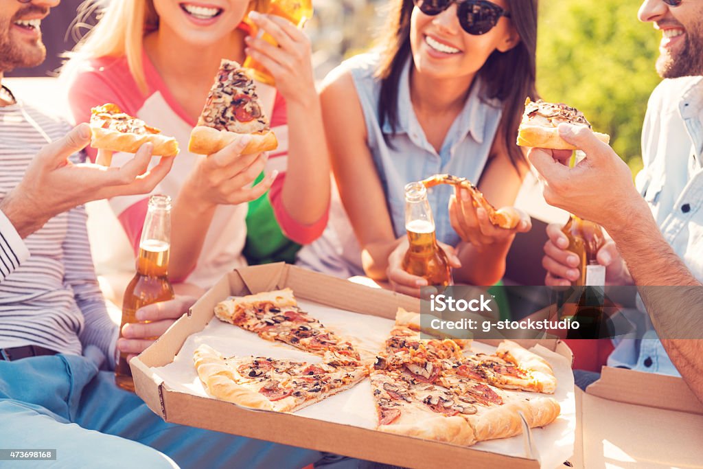 4 friends with drinks, sharing a pizza Close-up of four young cheerful people eating pizza and drinking beer outdoors 2015 Stock Photo