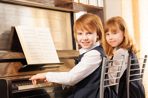 Cheerful beautiful small girls playing piano together with notes during lesson