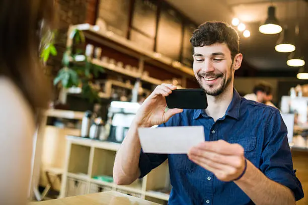 A smiling man takes a picture with his smart phone of a check or paycheck for digital electronic depositing, also known as "Remote Deposit Capture".  He sits across from someone at the coffee shop. Shot from over the womans shoulder, the focus on the man and check in the background. Horizontal image.