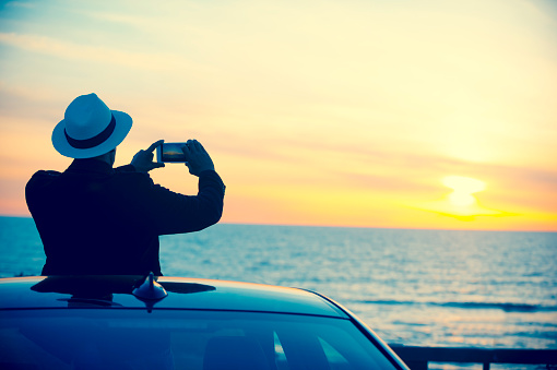 Man taking mobile phone picture at sunset. Mature age man on vacation taking a photograph of the sunset over the ocean. he is alone and wearing a hat and looking relaxed in a remote area. Sunset colours shot in California, USA