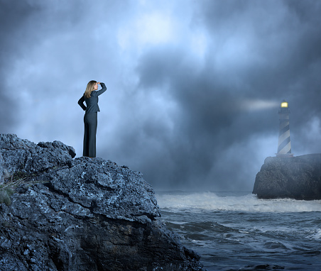 A businesswoman standing on a cliff above a rocky coastline as she looks through the fog and across the rough ocean waters towards a lighthouse high on a cliff in the distance. A beam of light emanates from the lighthouse through the fog out towards the ocean. Lots of negative space for copy.