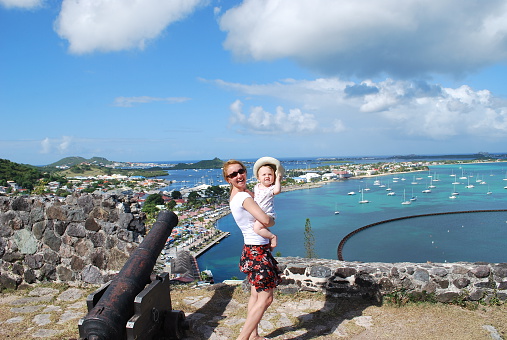 Mother and her 11 months daughter standing on a hill near historic Fort Saint-Louis in St. Marten Island. A view of Marigot and Anguilla Island on the background below as well as a luxury Marina Fort Louis that accommodates boats and yachts from all over the world.