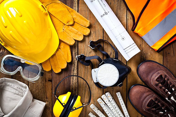 Personal safety workwear and construction blueprint shot directly above Personal protective workwear and blueprint shot directly from above on rustic wood background. The protective workwear includes hard hat, gloves, earmuff, goggles, steel toe shoes, and safety vest. Predominant colors: yellow and brown. DSRL studio photo taken with Canon EOS 5D Mk II and EF 100mm f/2.8L Macro IS USM safety equipment stock pictures, royalty-free photos & images