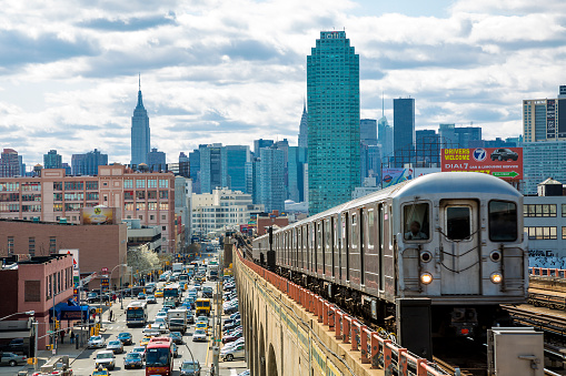 Train approaching  elevated subway station in Queens, New York. Finanancial buildings and New York skyline are seen in the background, on the left below can be seen a busy street full of cars at rush hour, cloudy, dramatic sky, horizontal orientation, USA