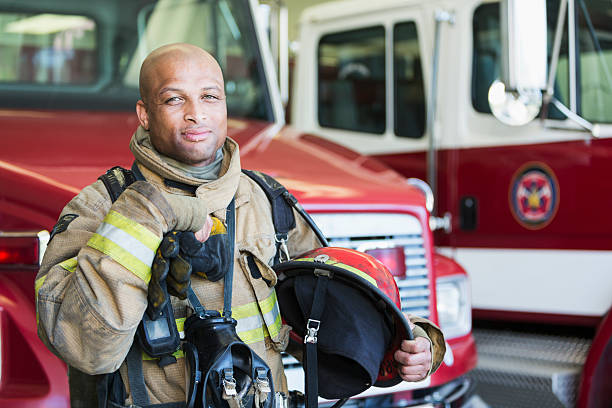 African American fireman at fire station Portrait of an African American fireman standing in front of a fire engine parked at the station.  He is serious and confident, wearing protective suit, holding gloves and a helmet.  He is ready to respond to an emergency. emergency services occupation stock pictures, royalty-free photos & images