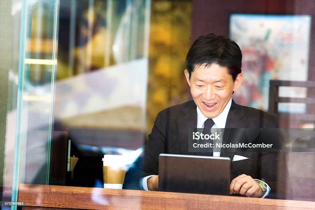 Happy Japanese businessman reads good news on tablet at cafe Happy Japanese businessman reads good news on tablet at a cafe. Shot through a window with urban reflections. His tablet is held by a keypad attachment. Cafe Stock Photo