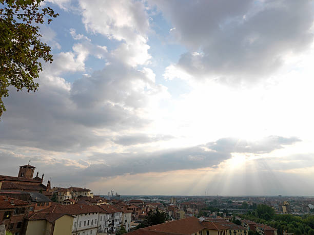 Looking across a Medieval town, over orange rooftops, towards the sun, clouds, and sun rays. Tree in foreground.