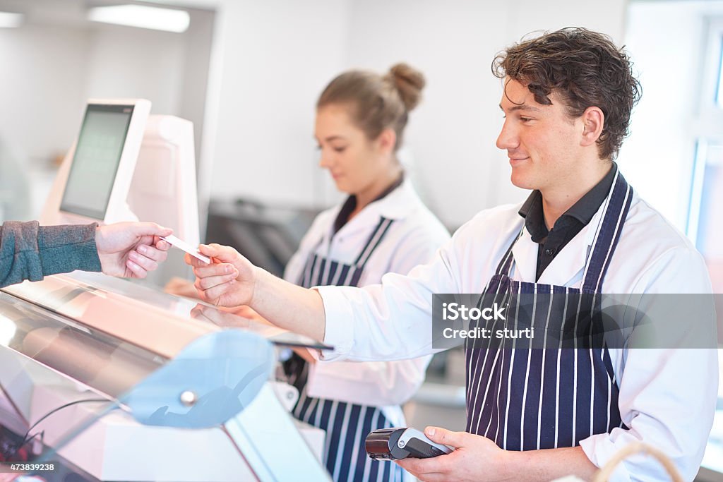 taking credit card payment in a butcher's shop A young male butcher apprentice is standing in front of the display fridge and smiling to a customer as he checks the credit card payment on the credit card reader  and hands back the card to him .In the background a young female apprentice is weighing some meat on electronic scales. The butchers are wearing white butcher's coats and blue striped aprons . Butcher's Shop Stock Photo