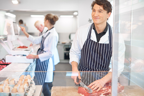 A young male butcher is standing at the butcher block slicing a piece of beef steak . He is looking up to chat to his customer .In the background a young female apprentice is weighing some meat on electronic scales. The butcher is wearing a white butcher's coat and blue striped apron .