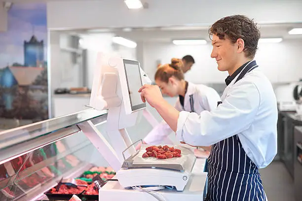 a young female butcher tends the meat counter in a butcher's shop. She is wearing a white coat and striped apron and is using electronic scales to weigh some diced steak . In the foreground a male colleague is seen weighing diced steak on some electronic scales .