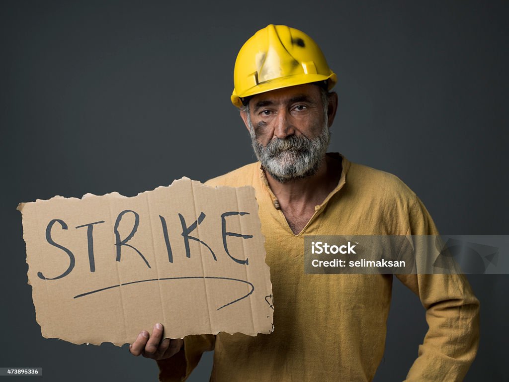 Senior Worker With Hardhat Holding Strike Placard Senior worker wearing a yellow hardhat and shirt holding a placard for strike.Word strike is written on placard.His face and hat is in dirt.He is a senior man and he has gray beard.Social security system and late retirement concept is aimed.The photo was shot with a medium format camera Hasselblad H4D in studio and in horizontal framing. 2015 Stock Photo