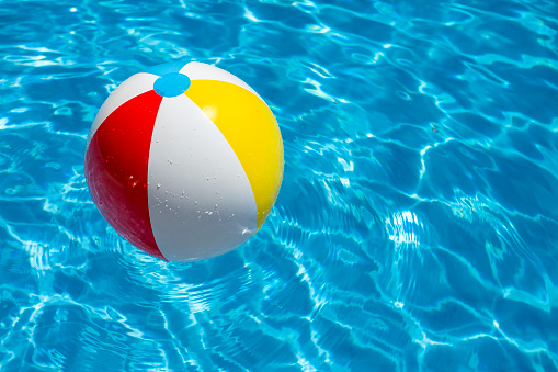 A multi colored beach ball floats on the water surface of a swimming pool. The wave pattern and the bright light blue color of the water gives a feeling of beach party and holiday. With copy space on the right.