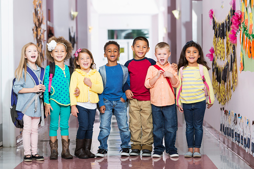 A multi-ethnic group of seven children standing in a row in a school hallway, laughing and smiling at the camera.  The little boys and girls are kindergarten or preschool age, 4 to 6 years.