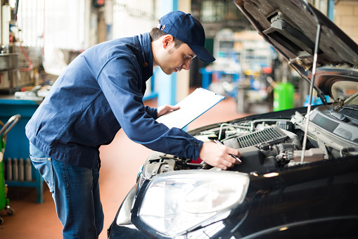 Close up view of hand of young male mechanic using laptop and his colleague with tablet, recording automobile engine checking collecting detailed information during his work on car workshop.