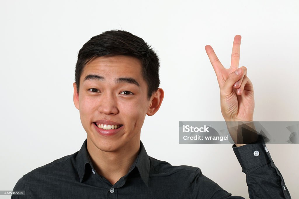 Smiling young Asian man giving the victory sign Smiling young Asian man giving the victory sign and looking at camera. 2015 Stock Photo